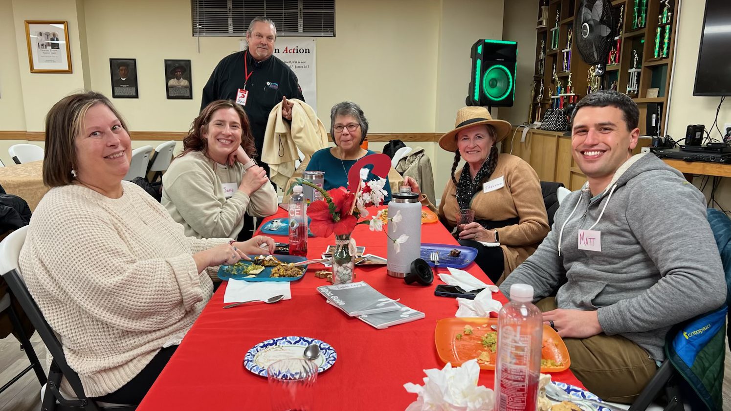 Six adults sit around a long red-table banquet, smiling and eating at a casual community gathering with a flower centerpiece and drinks nearby.