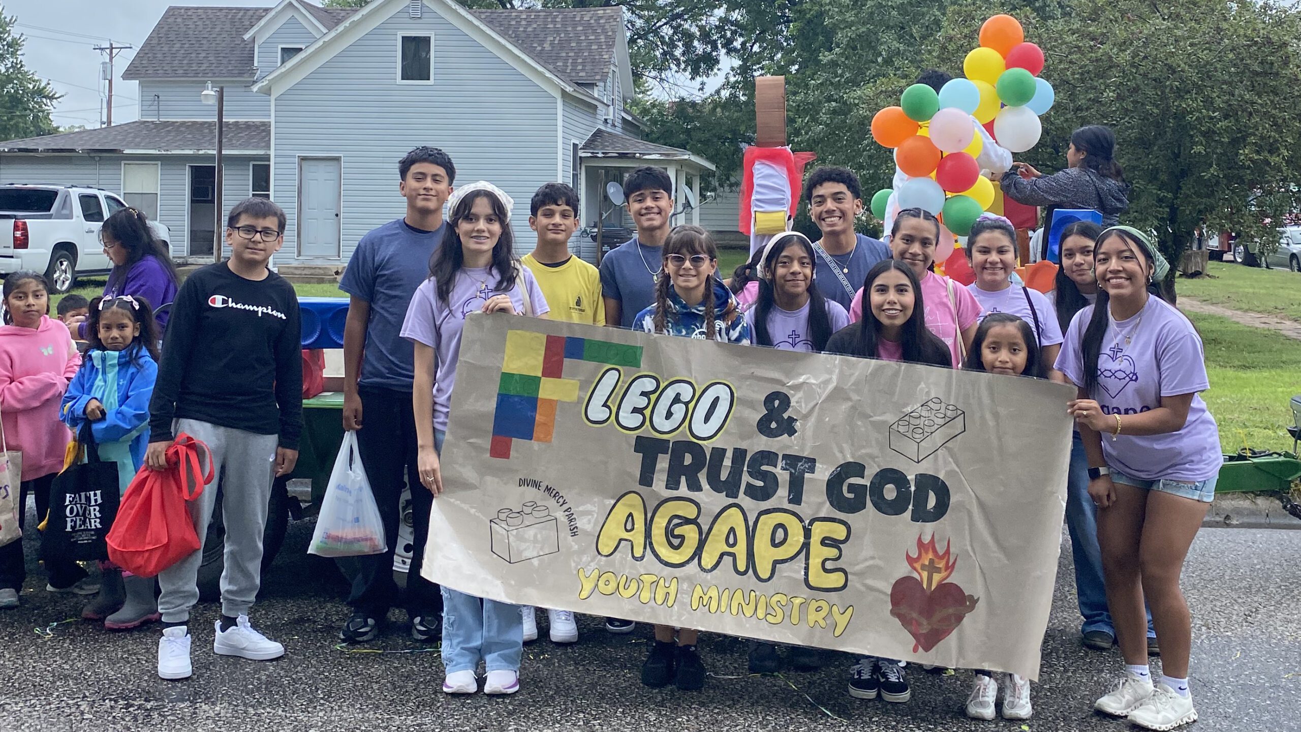 Group of smiling youths holding a banner for Agape Youth Ministry at a parade, with a colorful balloon arch in the background.