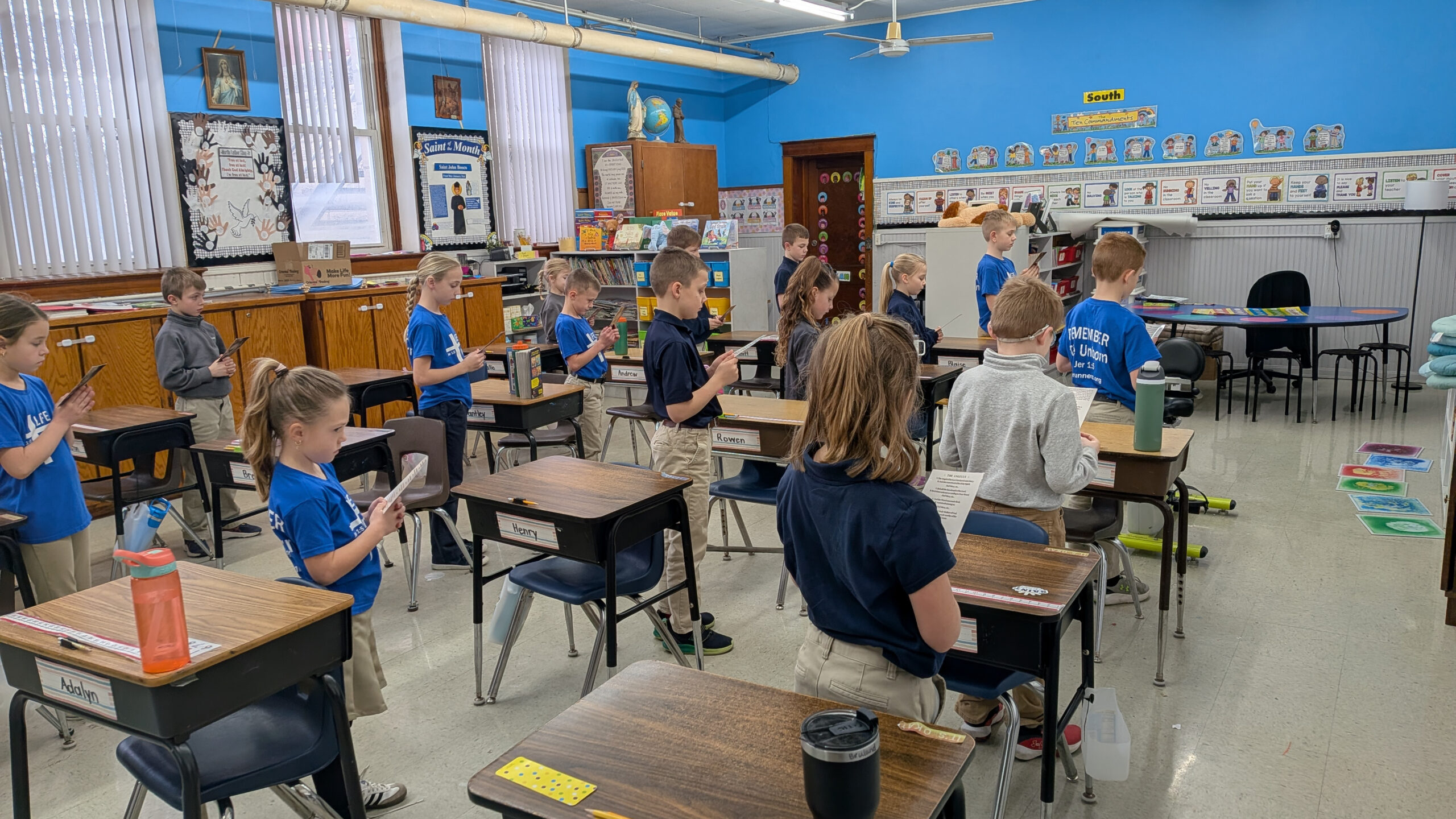 Students stand beside their desks in a bright blue classroom, looking at tablets or papers in unison for a lesson.
