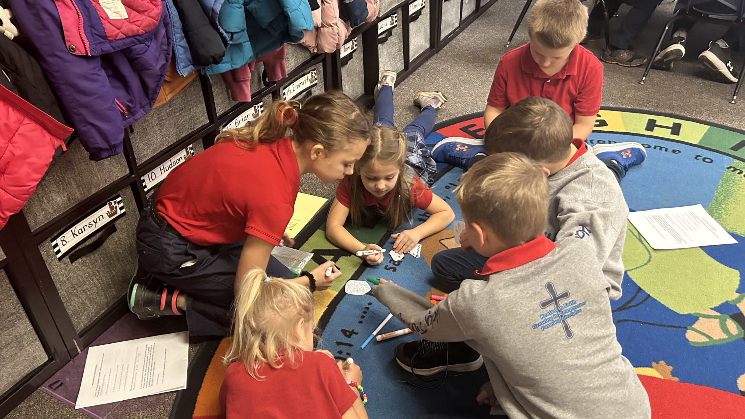 Group of children sitting on a colorful rug, drawing with markers while papers and worksheets are nearby in a classroom.