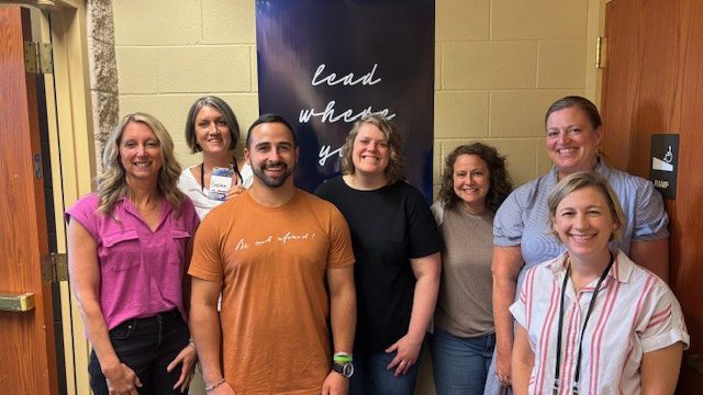 Group of seven adults smiling and posing for a photo in a hallway, with a dark banner behind that reads 'lead where you'.
