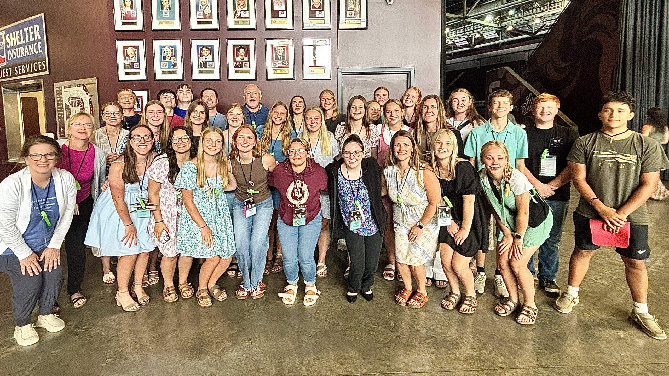 Group of about 30 people posing for a photo in a backstage area, wearing lanyards and casual summer clothes.