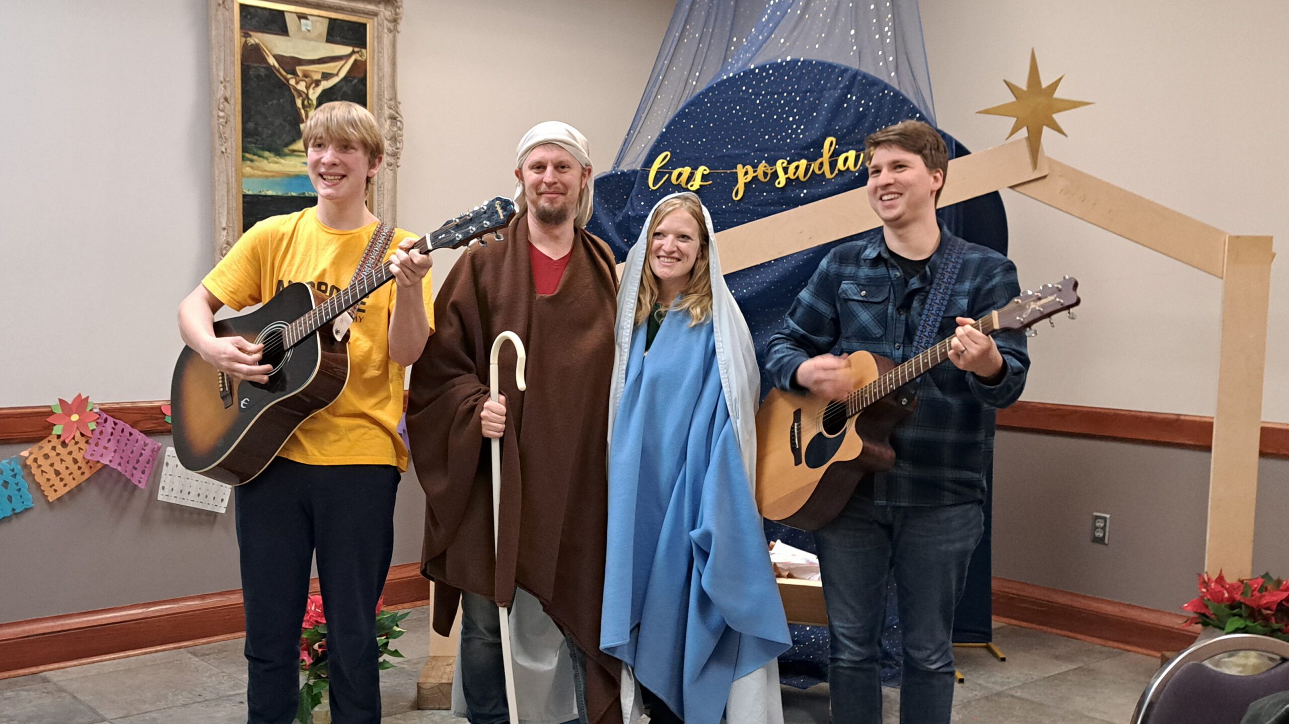 Group of four people in costumes posing with guitars and a staff, smiling on a small stage backdrop with a blue star backdrop and nativity scene theme.
