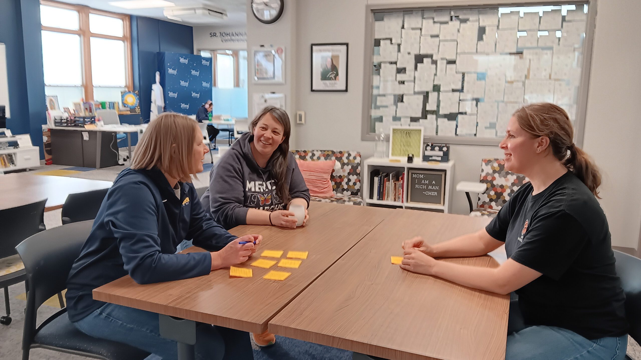 Three people sit around a table with yellow sticky notes, smiling and talking in a bright room.