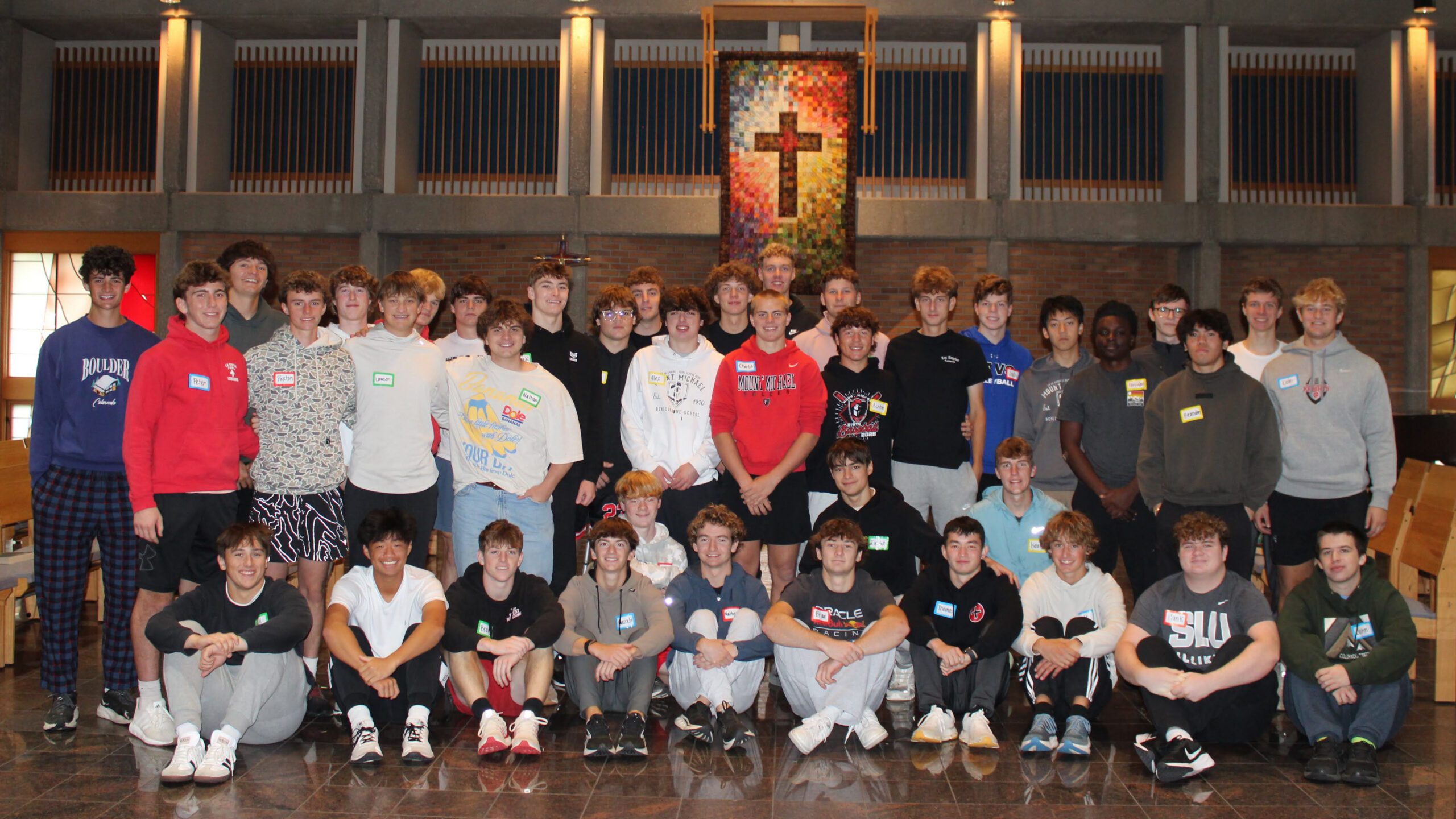 Large group of young people posing for a group photo in a hall, with a colorful cross banner hanging behind them.