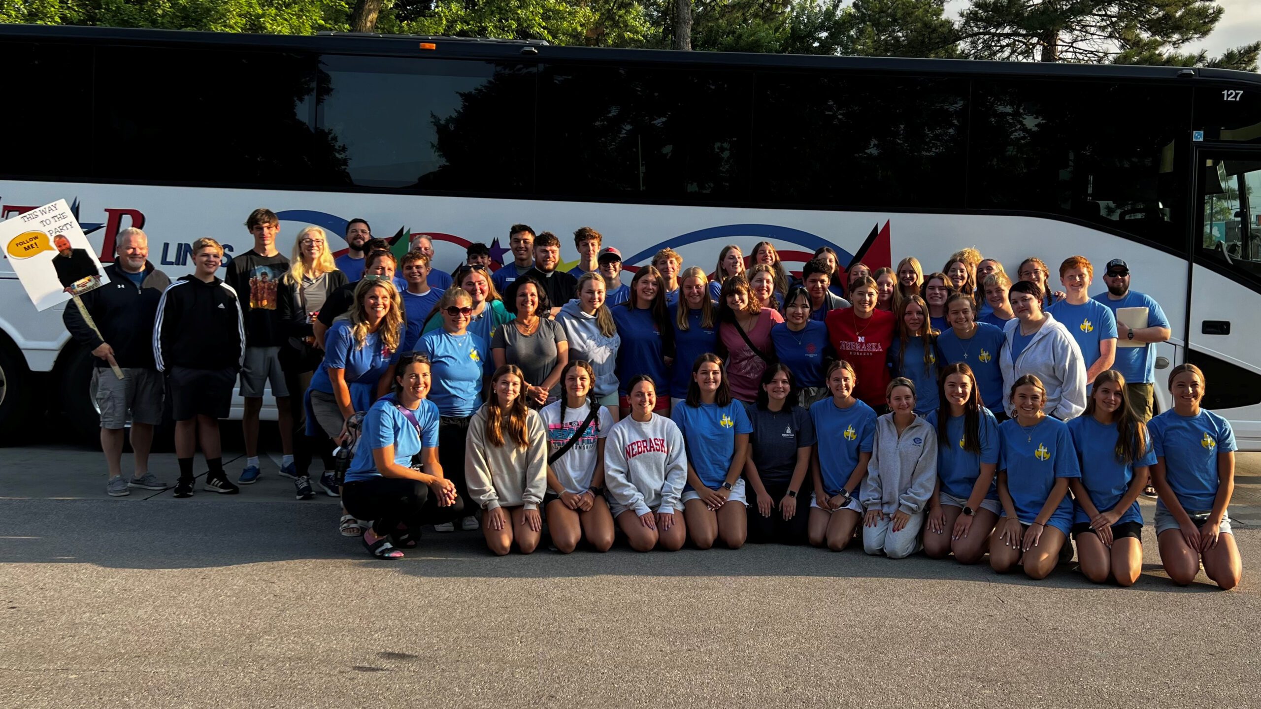 Large group of students posing in front of a white tour bus with blue/red graphics in the background.