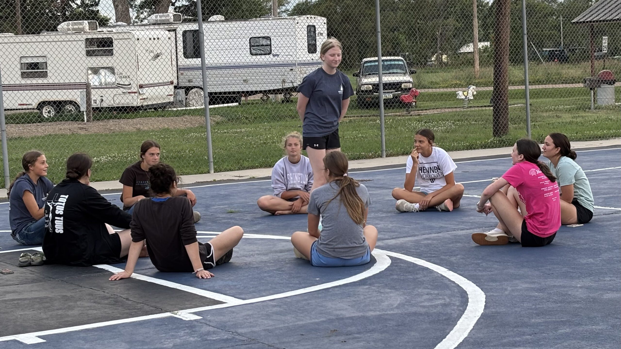 Group of young people sitting in a circle on a blue outdoor court, listening to a standing instructor.