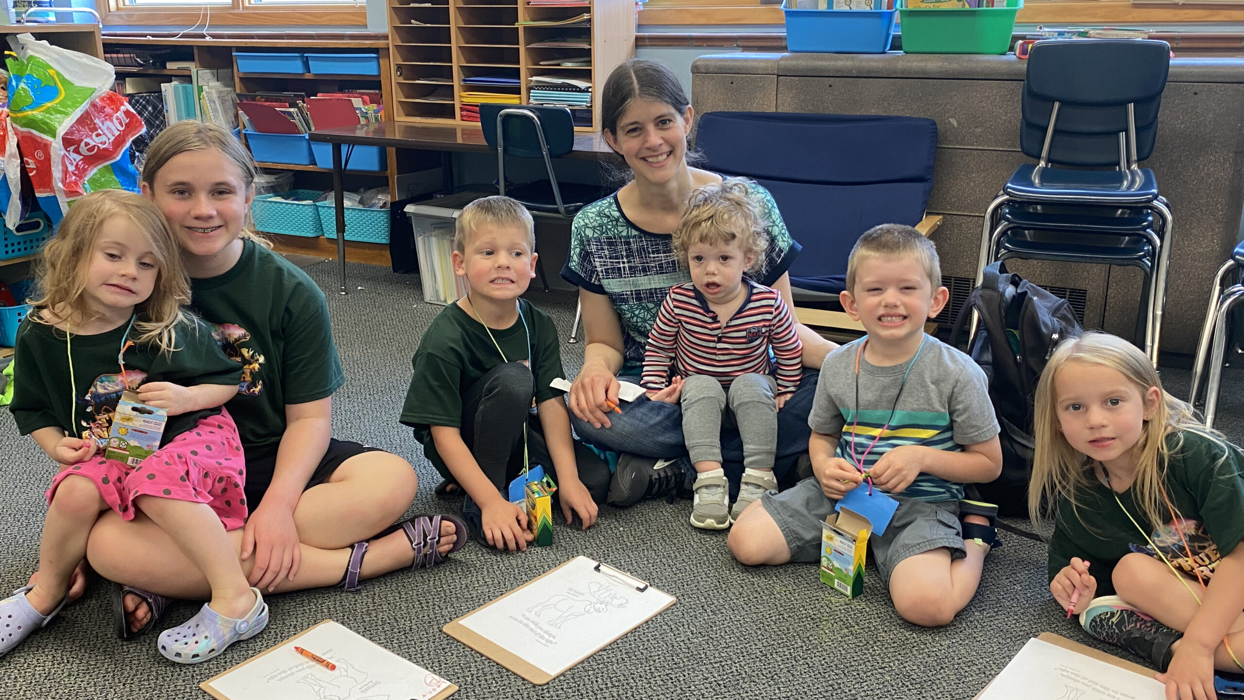 Smiling teacher and six children sit on a classroom carpet with clipboards and craft supplies during an activity.