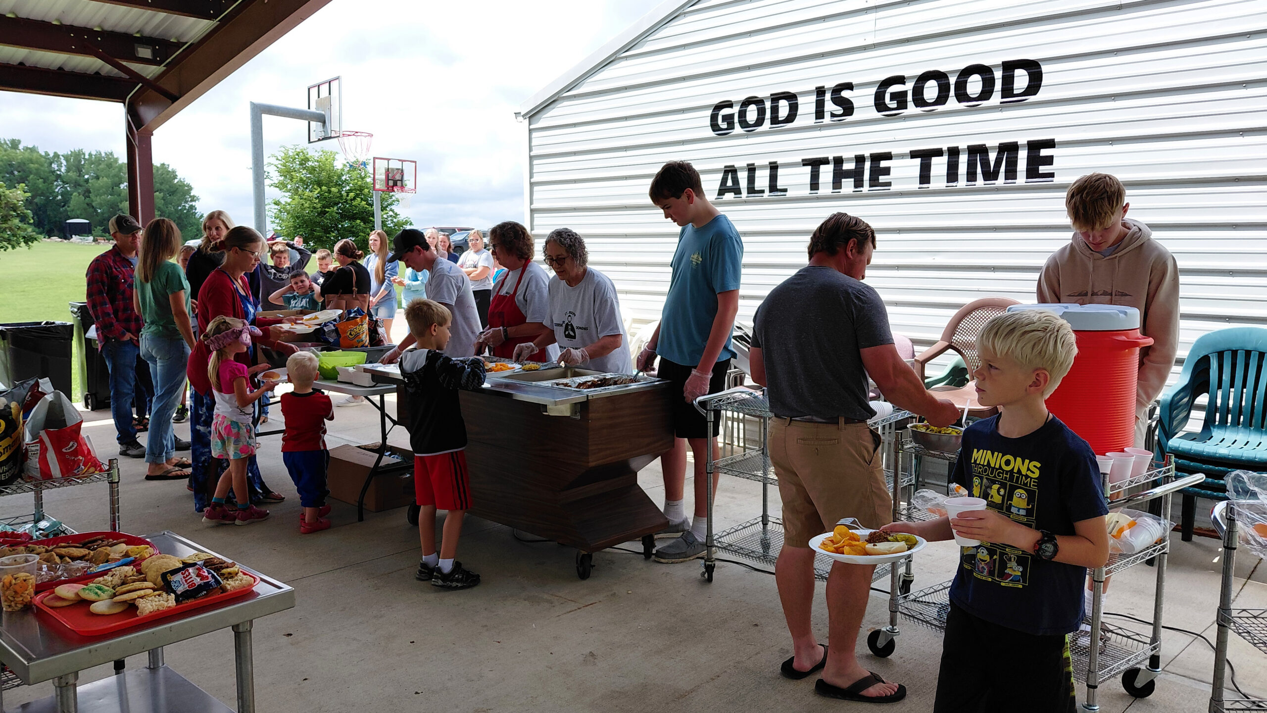 People line up at a community meal under a pavilion, volunteers serve food on a long table beside a metal building with the sign 'God Is Good All The Time'.