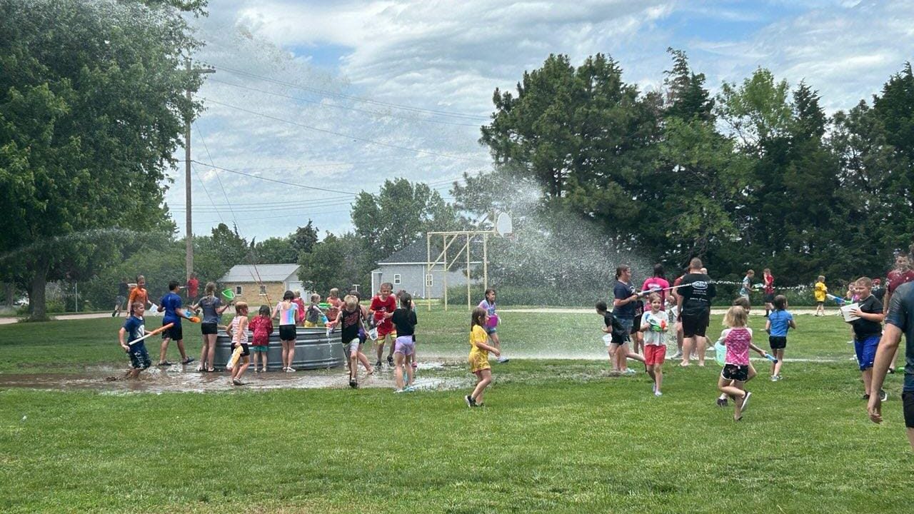Kids run and splash near a sprinkler and water buckets in a sunny park setting.