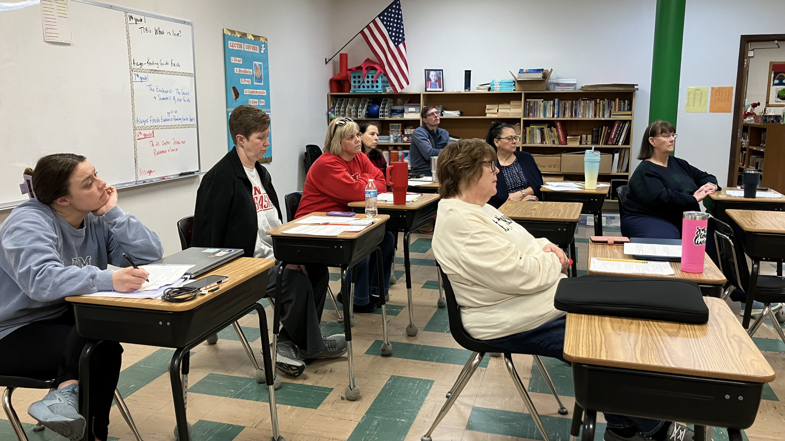 Adults seated at desks in a classroom, listening to a presentation; whiteboard, flag, and bookshelves visible in the background.