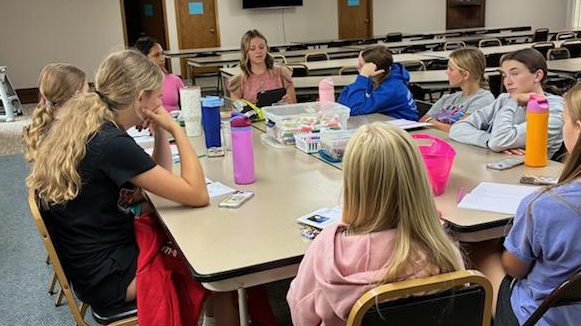 Group of students sitting around a classroom table while a teacher leads the discussion using a tablet at the head of the table.