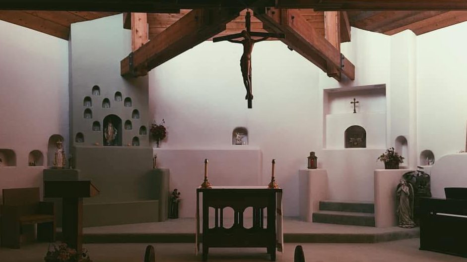 Chapel interior with a crucifix hanging over a simple wooden altar and candles, flanked by niche alcoves and statues on white walls.
