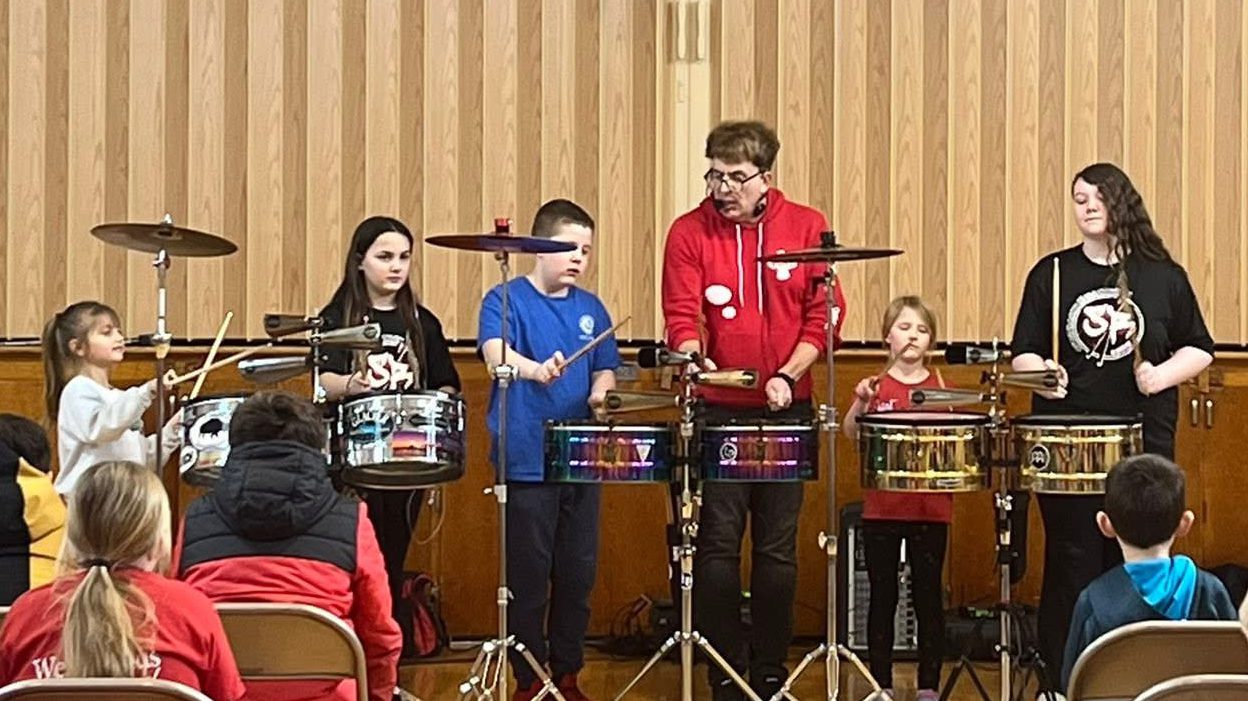 Instructor in a red hoodie leads a kids' drum ensemble on a wooden-stage backdrop with six students playing snare drums while a small audience watches.