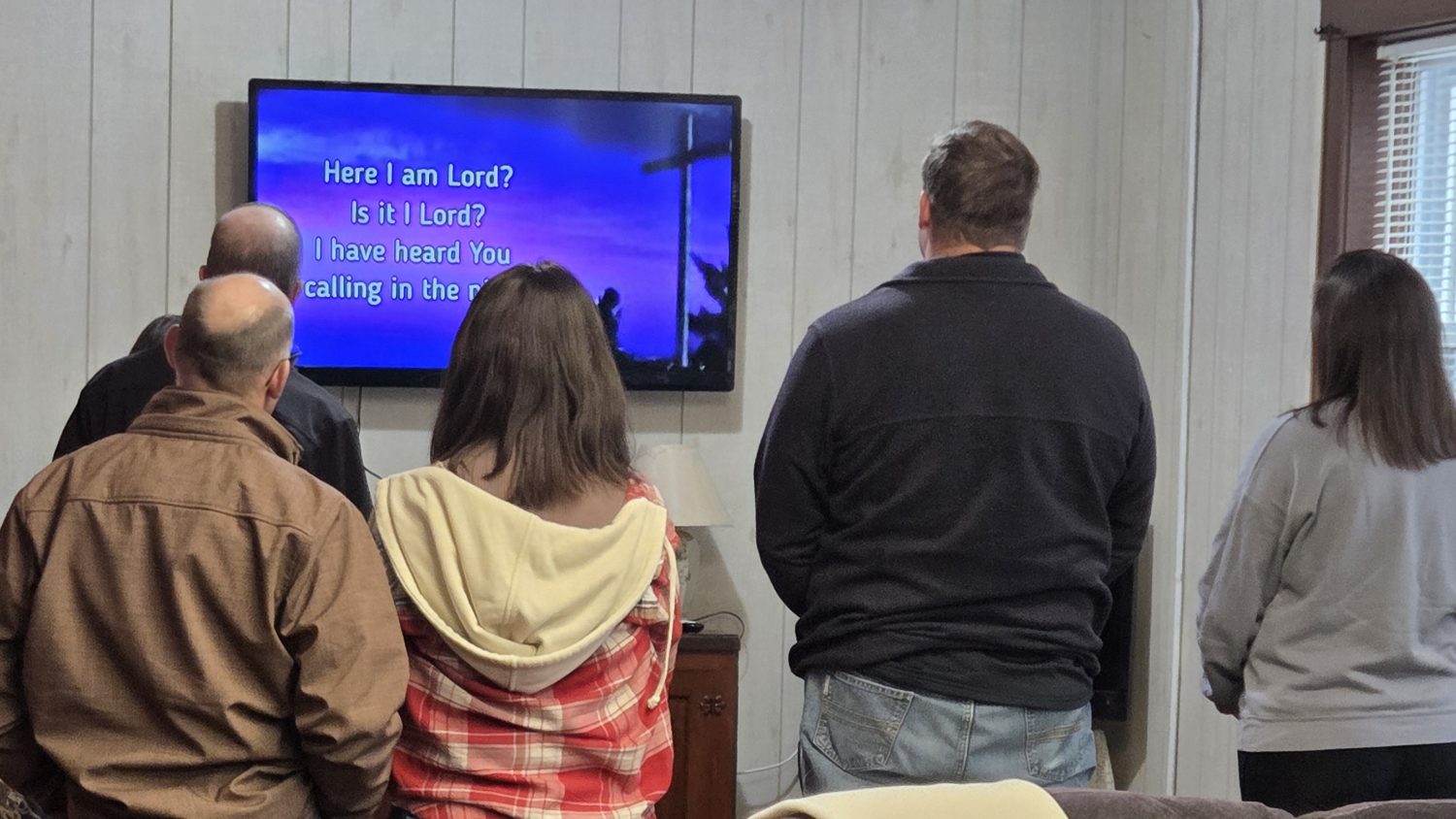 Group of adults standing in a room, facing a wall-mounted screen displaying worship lyrics.
