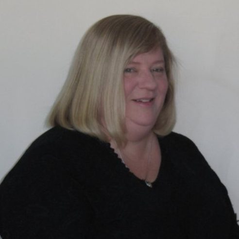 Smiling woman with shoulder-length blonde hair wearing a black top against a pale wall.
