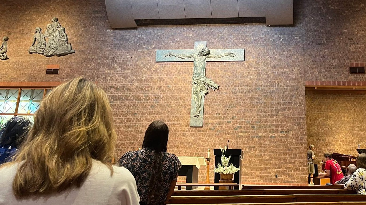 Interior of a church with a large crucifix on a brick wall, worshippers seated in pews facing the altar.Parabolic cross central.