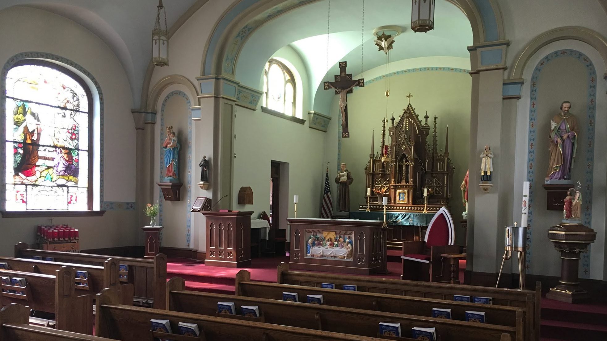 Interior of a church sanctuary with wooden pews, a red carpet aisle, and an ornate altar featuring a crucifix and statues against pale blue walls; stained glass window on the left adds colorful light.