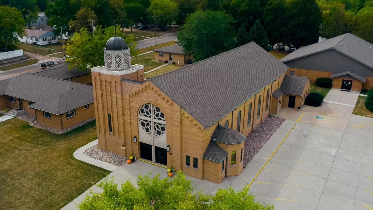 Aerial view of a brick church with a domed bell tower and a large circular stained-glass window above the entrance, surrounded by parking lots and green landscaping.