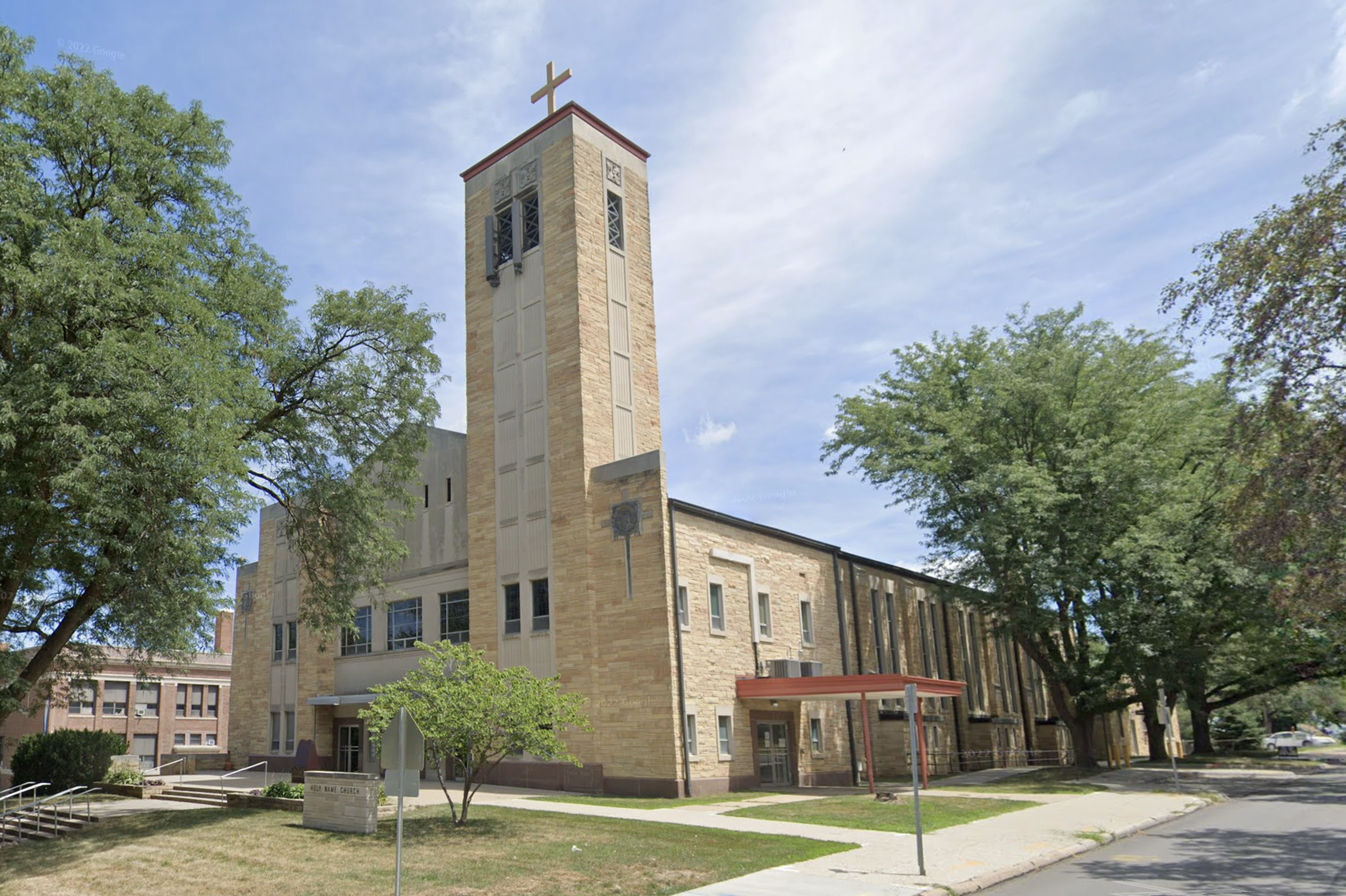 Brick church with a tall square bell tower topped by a cross, flanked by green trees on a sunny day