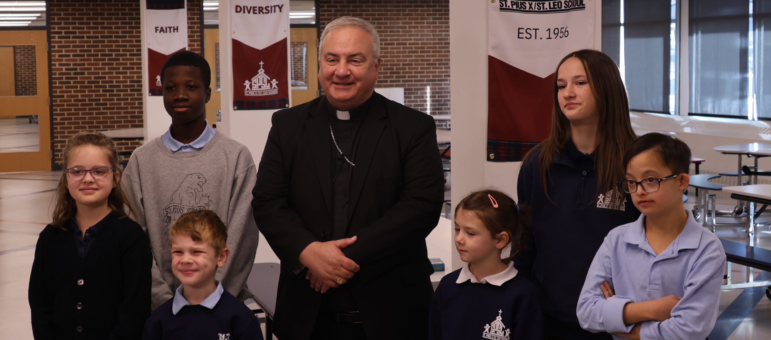 Priest in black clerical attire posing with several students in a school hallway with banners in the background.